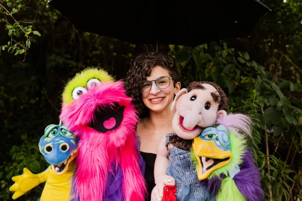 Tansy smiling with her ventriloquist puppets; Molly the Monster, Cletus the Redneck, Gus the Magical Dragon, and Heidi the Non-binary Bird